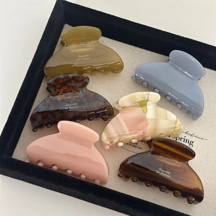 Six large hair claw clips in different colors—yellow, blue, tortoiseshell, pink, cream, and brown—are displayed on a beige fabric tray.