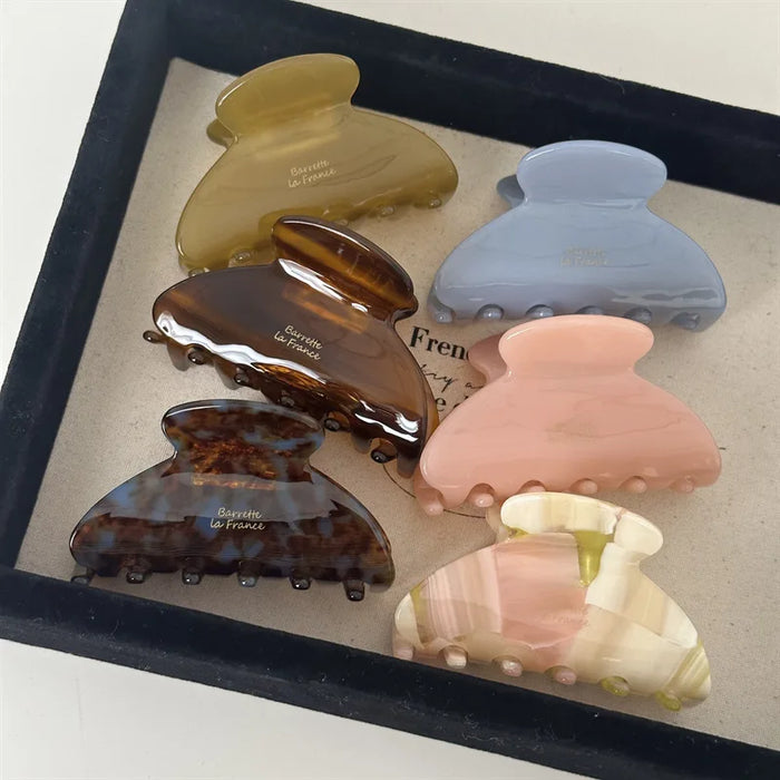 Six large hair claw clips in different colors—yellow, blue, tortoiseshell, pink, cream, and brown—are displayed on a beige fabric tray.